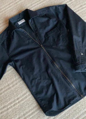 John Henric Full Zip - Stilren svart overshirt från John Henric med klassisk krage, bröstficka och hel dragkedja framtill. Tillverkad i bomull för en skön känsla och passar perfekt som lager över t-shirt eller skjorta. Enkel och clean design som funkar till många outfits.