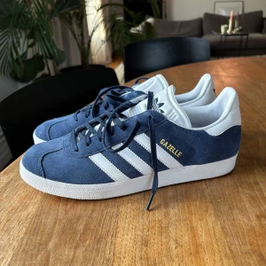 Adidas Gazelle blå/vita sneakers - Säljer ett par klassiska Adidas Gazelle sneakers i marinblå mocka med vita detaljer och de ikoniska tre ränderna på sidan. Skorna har snörning, vit sula och Adidaslogga på hälen. Knappt använda 