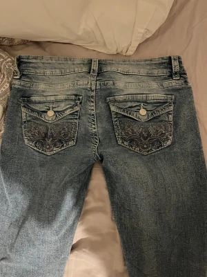 Blå, lågmidjade byxor - Snygga ljusblå jeans från Gina Tricot med slitningar framtill och detaljerade broderier på bakfickorna. Jeansen har normal passform, uppsprättade längst ner för att bli något längre.  Storlek M 14+