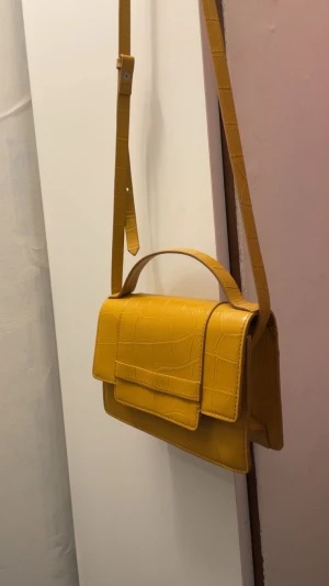 Gul axelväska med krokopräglat mönster - Snygg gul axelväska med krokopräglat mönster och justerbar rem. Väskan har en stilren, rektangulär form och ett praktiskt lock framtill. Materialet är syntet med en glansig finish som ger en cool vibe. Perfekt för att lyfta din outfit med lite färg. Aldrig använt tidigare!
