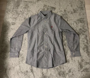Grå skjorta från Polo Ralph Lauren - Snygg grå skjorta från Polo Ralph Lauren med klassisk krage och knappar framtill. Skjortan har lång ärm och ikonisk röd logobrodering på bröstet. Tillverkad i mjuk bomull som känns skön mot huden. Perfekt för dig som gillar stilrena och tidlösa plagg.