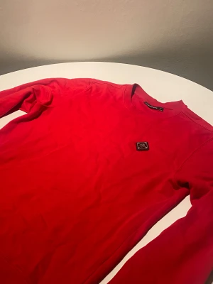 Röd sweatshirt från J.Lindeberg - Riktigt snygg röd sweatshirt från J.Lindeberg med metallpatch på bröstet. Tröjan har rund hals och långa ärmar, tillverkad i mjuk bomull. Perfekt för dig som gillar stilrena och sportiga plagg med en cool detalj.