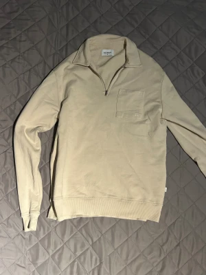 Beige half zip tröja från Woodbird - Snygg beige långärmad tröja från Woodbird med half zip och krage. Tröjan har en bröstficka med diskret broderad logga och ribbade muddar vid ärmslut och nederkant. Perfekt för dig som gillar stilren och enkel design.