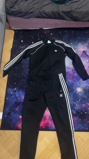 Svarta Adidas mjukisbyxor med vita ränder - Svarta mjukisbyxor från Adidas med klassiska vita ränder längs sidorna och Adidas-logga på benet. Byxorna har elastisk midja och raka ben, tillverkade i mjukt syntetmaterial som är skönt att ha på sig. Perfekta för chill eller träning.