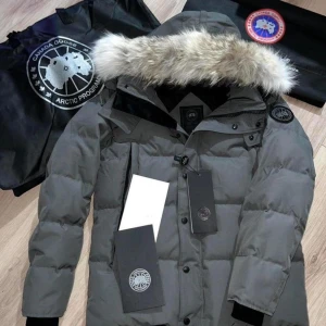 Grå dunjacka från Canada Goose - Säljer en grå dunjacka från Canada Goose med pälskantad huva och klassisk logga på ärmen. Jackan har knappar och dragkedja framtill, quiltad design och lång ärm. Perfekt för kalla dagar och riktigt snygg streetstil.