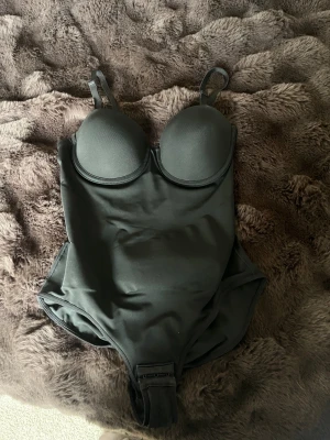 Svart body  med bygel och justerbara axelband - Säljer en svart body med bygel och vadderade kupor. Den har justerbara axelband och en hakknäppning i grenen. Materialet är mjukt och stretchigt, perfekt för strand eller pool. Snygg söm på baksidan för extra passform.