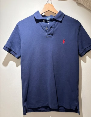 Ralph lauren pike S - Snygg marinblå pikétröja från Polo Ralph Lauren med röd broderad logga på bröstet. Tröjan har korta ärmar, klassisk krage och två knappar framtill. Slim fit-modell i mjuk bomull som sitter riktigt bra och känns fräsch till jeans eller shorts.