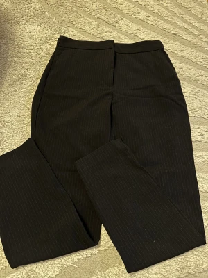 Svarta kostymbyxor med diskreta ränder - Snygga svarta kostymbyxor från Zara med diskret randigt mönster. Klassisk rak passform, hög midja och praktiska fickor bak. Stängs med knapp och dragkedja framtill. Perfekta för en stilren look.