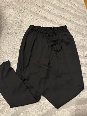 Svarta kostymbyxor med snörning - Svarta kostymbyxor med elastisk midja och snörning framtill. Byxorna har raka ben och en bakficka. Perfekta för chill dagar och har en enkel, stilren look. Materialet känns mjukt och bekvämt mot huden.