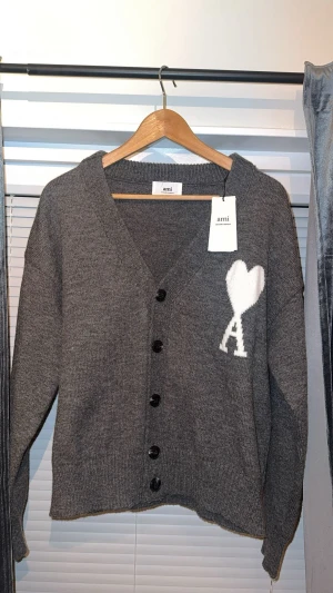 Grå kofta från Ami med hjärtdetalj - Grå stickad kofta från Ami Alexandre Mattiussi med svart knäppning framtill och vit hjärtdetalj med bokstaven A på bröstet. Koftan har v-ringad hals, ribbade muddar och långärmad passform. Perfekt för dig som gillar stilrena och coola detaljer.