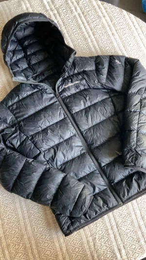 Dunjacka Peak Performance - Svart dunjacka från Peak Performance med huva och dragkedja framtill. Jackan har quiltad design och logotyp på bröstet. Perfekt för kalla dagar och har en snygg, normal passform som funkar till streetwear. Materialet är syntet och dun. Inga hål/defekts.