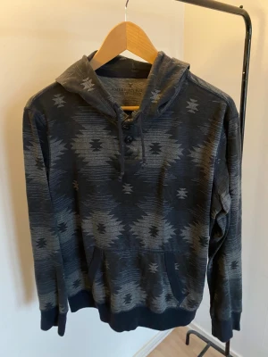 Mörkgrå hoodie American Eagle -storlek extra small - Snygg luftig mörkgrå hoodie från American Eagle Outfitters i storlek XS. Tröjan har ett unikt mönster, huva med snörning och knappar vid halsen. Tillverkad i mjuk bomull och polyesterblandning, perfekt för chill dagar. Fickor framtill och långa ärmar.