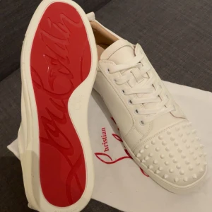 Christian Louboutin vita sneakers med nitar - Exklusiva vita sneakers från Christian Louboutin i skinn med ikonisk röd sula och coola nitar på tån. Skorna har snörning och en stilren, modern look som sticker ut. Perfekta för dig som vill ha något unikt och lyxigt i garderoben.