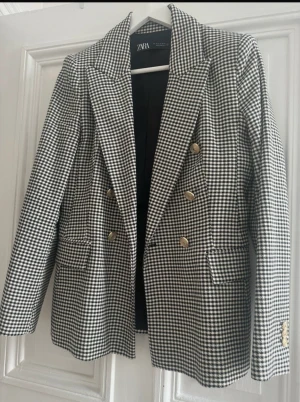 Rutig svartvit kavaj från Zara - Snygg svartvit rutig kavaj från Zara med dubbelknäppning och guldiga knappar. Klassisk modell med bred krage och fickor framtill. Perfekt för dig som vill ha en stilren och trendig look. Materialet är mjukt och följsamt, troligtvis polyesterblandning.