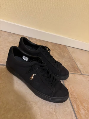 Svarta sneakers från Polo Ralph Lauren - Svarta sneakers från Polo Ralph Lauren med klassisk låg profil och rund tå. Skorna har svart textilöverdel, svarta snören och Polo-logga broderad i guld på sidan. Sulan är brun undertill med Polo-tryck. Perfekta för en clean och stilren look.