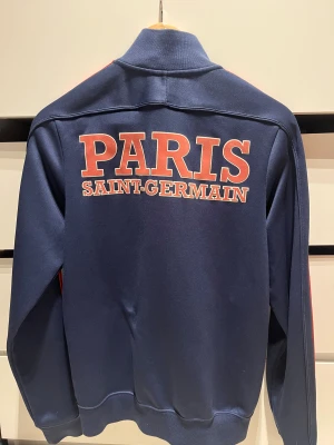 PSG träningströja marinblå S - Paris Saint-Germain ziptröja från Nike i marinblått med röda detaljer längs ärmarna och PSG-logga på bröstet. Tröjan har hög krage, dragkedja framtill och fickor med dragkedja. Nike paris saint germain