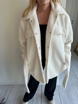 Vit teddyjacka med bälte - Säljer en fluffig vit teddyjacka med bred krage, stora bröstfickor. Jackan har ett matchande bälte i midjan och långa ärmar. Perfekt för dig som gillar en chill och cozy stil under kyliga dagar. 😍