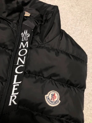 Svart dunväst från Moncler, storlek L - Svart dunväst från Moncler med quiltad design och hög krage. Västen har tydlig Moncler-logga på bröstet och vit broderad Moncler-text längs dragkedjan. Två sidofickor med dragkedja och coolt serietryck på insidan. Perfekt för lager-på-lager och streetstyle.