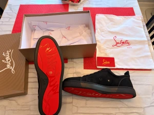Svarta sneakers från Christian Louboutin - Snygga svarta sneakers från Christian Louboutin med klassisk röd sula och diskret logga. Skorna har rund tå, platt sula och är tillverkade i mocka med svarta snören. Kommer med originalkartong och dustbag. Perfekt för dig som gillar exklusiv streetstyle.