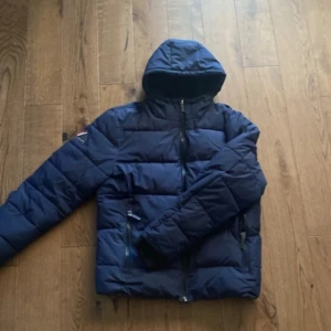 Mörkblå pufferjacka från Superdry - Säljer en mörkblå pufferjacka från Superdry med huva och broderad logga på ärmen. Jackan har dragkedja framtill, två sidofickor med dragkedja och mjukt fleecefoder på insidan. Perfekt för kalla dagar och har en clean, sportig look.
