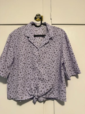 Ljuslila blommig skjorta från H&M - Superfin ljuslila skjorta från H&M med små bruna blommor över hela tyget. Skjortan har korta ärmar, knappar framtill och en snygg knytning längst ner. Perfekt för dig som gillar en chill och söt stil.