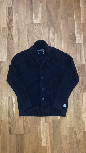 Svart stickad Cardigan från Jack & Jones - Mysig svart stickad kofta från Jack & Jones med grov ribbad struktur och klassisk krage. Koftan har knappar framtill och en liten etikett på nederkanten. Perfekt för dig som gillar stilrena och bekväma plagg. Storlek M och passar mig som är 175cm perfekt! Vid fler frågor eller funderingar tveka inte på att skriva!