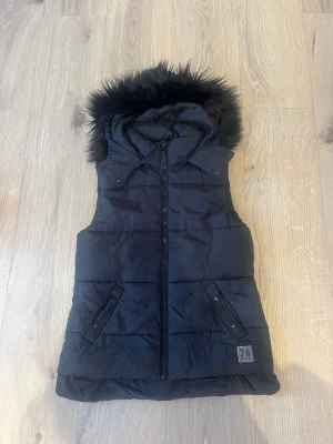 Svart dunväst med fuskpäls från H&M strl 146/152 - Svart dunväst från H&M med huva och fuskpälsdetalj runt kanten. Västen har dragkedja framtill, två snedställda fickor med knappar och quiltad design. Perfekt för lager-på-lager och snygg till streetwear.