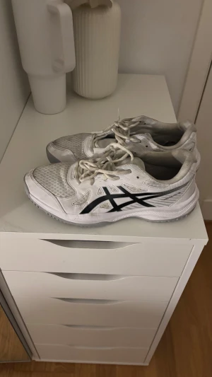 Vita Asics Upcourt 6 sneakers - Fräscha vita Asics Upcourt 6 sneakers med svart logga på sidan. Skorna har meshpaneler för extra ventilation och snörning framtill. Snygg sportig design med rund tå och platt sula, perfekta för träning eller häng. Materialet är syntet och mesh.