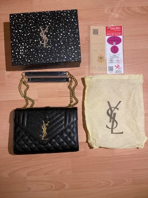 Svart quiltad YSL axelväska i skinn - Snygg svart axelväska från YSL i quiltat skinn med guldiga kedjor och ikoniskt YSL-emblem framtill. Väskan har kuvertformad front, magnetlås och ett fack inuti. Perfekt för dig som vill ha en lyxig och stilren accessoar med exklusiv känsla.