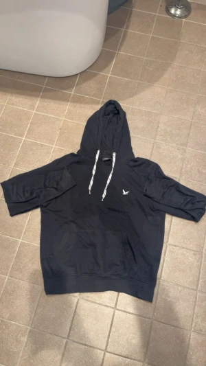 Svart hoodie från Blend med snörning - Svart hoodie från Blend med vit snörning i huvan och en liten logga på bröstet. Tröjan har en klassisk känguruficka framtill och lång ärm. Perfekt för chill dagar och enkel att matcha med jeans eller joggers.