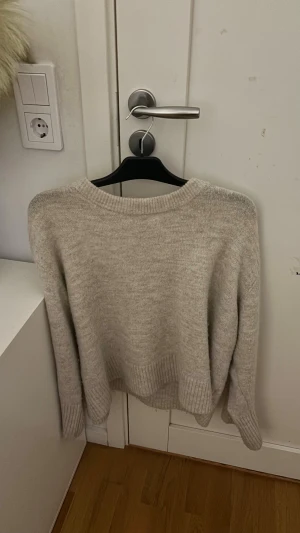 Beige stickad tröja med rund hals - Mysig beige stickad tröja med rund hals och ribbade muddar vid ärmslut och nederkant. Perfekt för kyliga dagar och enkel att matcha med jeans eller kjol. Tröjan har en loose passform och långa ärmar för extra komfort.