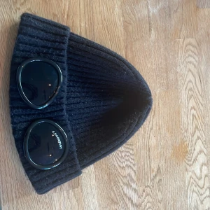 Svart ribbad mössa C.P. Company - Svart ribbstickad mössa från C.P. Company med ikoniska svarta glasögon-linser på uppviket. Mössan är gjord i mjuk ull och har en clean, urban look som sticker ut. Perfekt för dig som gillar streetwear och vill ha något unikt på huvudet. Givetvis äkta!