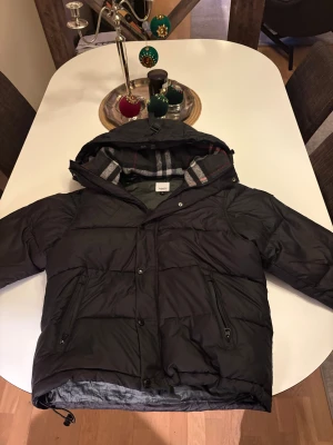 Svart pufferjacka från Burberry - Svart pufferjacka från Burberry med avtagbara ärmar så den kan bäras som väst. Jackan har huva, dragkedja och knappar framtill, samt två fickor med dragkedja. Insidan av huvan har klassiskt rutigt Burberry-mönster i grått, vitt och rött. Dragkedjan kan krångla lite då och då men det är inget problem