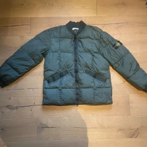 Grön pufferjacka från Stone Island - Snygg grön pufferjacka från Stone Island med ikonisk patch på vänster ärm. Jackan har ribbad krage, två snedställda fickor och dragkedja framtill. Materialet är polyester och jackan är quiltad för extra värme. Perfekt för dig som gillar streetwear och vill sticka ut.