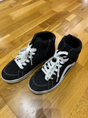 Svarta high-top sneakers med vit sula - Säljer ett par svarta high-top sneakers från Vans med vit sula och vita snören. Skorna har klassisk rund tå, kontrastsömmar och den ikoniska vita sidoremsan. Materialet är slitstark canvas och de har en platt sula som ger en skön streetstil.