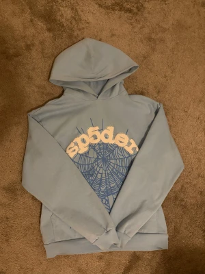 Ljusblå hoodie från Sp5der - Säljer en ljusblå hoodie från Sp5der med stor spindelnätsgrafik och broderad logga i vitt på bröstet. Tröjan har huva och långa ärmar, tillverkad i mjuk bomull som är skön att ha på sig. Perfekt för dig som gillar streetwear och coola prints.