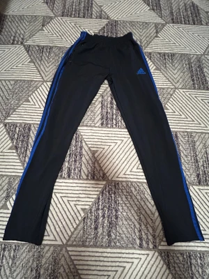 Svarta Adidas träningsbyxor med blå ränder - Snygga svarta träningsbyxor från Adidas i storlek L med blå ränder längs sidorna och Adidas-logga på låret. Byxorna har elastisk midja och dragkedja vid fickan. Perfekta för sport eller chill.