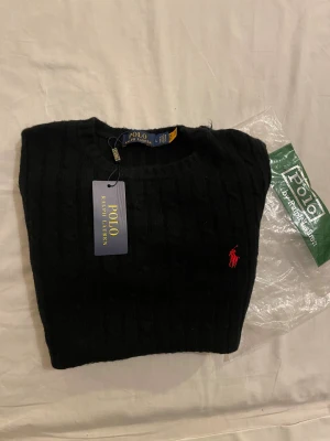 Svart stickad tröja Polo Ralph Lauren - Svart stickad tröja från Polo Ralph Lauren med klassisk kabelstickad design och röd broderad logga på bröstet. Rund halsringning och långa ärmar. Tröjan är gjord i mjuk ull och har en ribbad kant vid ärmslut och nederkant. Den är helt ny och passade inte för när jag bästelde den från Ralph Lauren i L men passar S.
