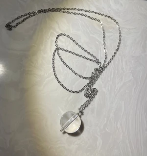 Halsband med glaskula och silverkedja - Unikt halsband med en rund, transparent glaskula som hänge. Kedjan är i silverfärgad metall och har en enkel, stilren design. Glaskulan är genombruten av en tunn metallstav som ger ett modernt och minimalistiskt intryck. Perfekt för dig som gillar stilrena accessoarer.