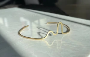 Guldigt minimalistiskt armband vågform - Unikt armband i guldig mässing med en cool vågformad design. Öppen modell som enkelt kan justeras runt handleden. Perfekt för dig som gillar stilrena och moderna accessoarer med en twist.