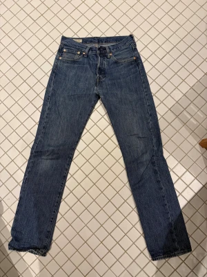 Levi's 501 blå jeans straight fit i storlek 28:32 - Klassiska Levi's 501 jeans i blå tvätt med raka ben och fem fickor. Jeansen har normal passform och är tillverkade i slitstarkt bomullsjeansmaterial. De har knappgylf och detaljerade sömmar, perfekt för en chill och tidlös look.