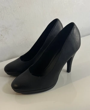 Svarta pumps från Graceland - Klassiska svarta pumps från Graceland med rund tå och hög klack. Skorna har en diskret struktur i materialet som ger en snygg touch. Perfekta för dig som gillar stilrena och tidlösa skor. Tillverkade i syntetmaterial.