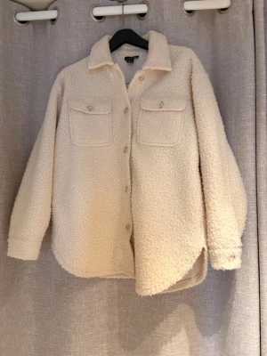 Krämvit teddyjacka - Mysig krämvit teddyjacka med två bröstfickor och knäppning framtill. Jackan har klassisk skjortkrage och är tillverkad i fluffigt teddy-material.