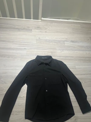 Svart overshirt från Zara - Stilren svart overshirt från Zara med klassisk krage, två stora bröstfickor och knäppning framtill. Skjortan har lång ärm och en rak, något loose passform. Perfekt att slänga över en t-shirt för en clean look.