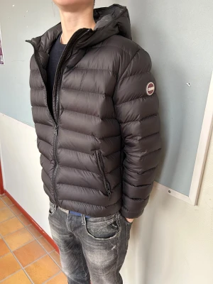 Svart dunjacka från Colmar - Säljer en svart dunjacka från Colmar med huva och snygga horisontella sömmar. Jackan har dragkedja framtill, två sidofickor med dragkedja och Colmar-logga på ärmen. Perfekt för kalla dagar och riktigt snygg stil.