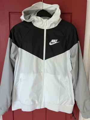 Nike svartvit vindjacka XL - Nike vindjacka i svart och vitt med huva och hel dragkedja. Jackan har två fickor med dragkedja och meshfoder på insidan. Snygg sportig look med klassisk Nike-logga på bröstet. Passar dig som gillar streetwear och sportig stil.