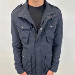 Fieldjacka från Woolrich - Mörkblå fieldjacka från Woolrich med flera fickor framtill, axelklaffar och hög krage. Jackan har både dragkedja och knappar. Jackan har aldrig använts så är i perfekt skick. Nypris är runt 6000kr. Snygg och funktionell design med normal passform. Perfekta julklappen till din man, kompis eller son.