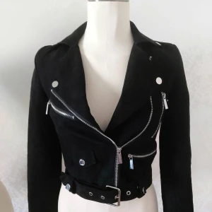 Svart croppad jacka med bälte - Snygg svart croppad jacka med flera silvriga dragkedjor och knappar. Jackan har ett brett bälte i midjan och klassisk krage. Perfekt för dig som gillar edgy stil och vill sticka ut med coola detaljer.