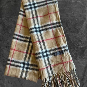 Rutig beige halsduk från Burberry - Snygg klassisk halsduk från Burberry  beige med rutigt mönster i svart, vitt och rött. Gjord av 100% lammull och har fransar i ändarna. Perfekt accessoar för att lyfta din outfit med ikonisk brittisk stil.