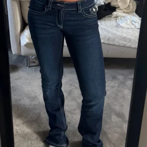 Mörkblå bootcut jeans med kontrastsömmar - Säljer ett par mörkblå lowwaist jeans med bootcut passform och vita kontrastsömmar. Jeansen har klassiska fickor fram och bak, bakfickorna har lock och knapp. 